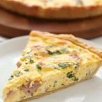 A slice of Tortilla Egg Bake on a white plate, with the remaining quiche on a wooden board in the background. The golden, flaky crust and creamy, cheesy filling are garnished with fresh herbs.