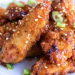 Golden brown crispy chicken tenders coated in a sticky honey garlic glaze, garnished with sesame seeds and sliced green onions, served on a white plate.