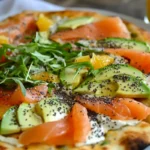 A gourmet pizza topped with smoked salmon, avocado slices, melted cheese, fresh herbs, and chia seeds, served on a white plate with a glass of orange juice.