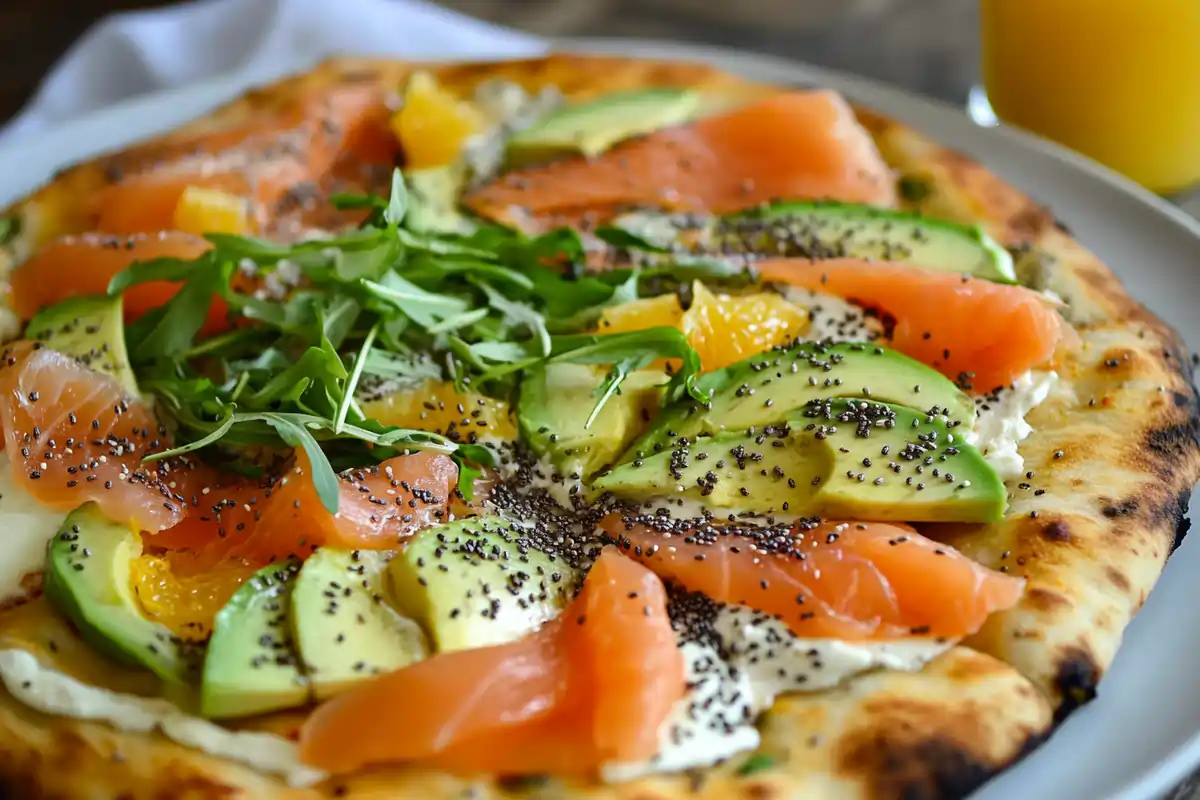 A gourmet pizza topped with smoked salmon, avocado slices, melted cheese, fresh herbs, and chia seeds, served on a white plate with a glass of orange juice.