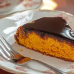 a piece of brazilian carrot cake taken by a fork, the rest of the cake is placed in a white plate on the table.