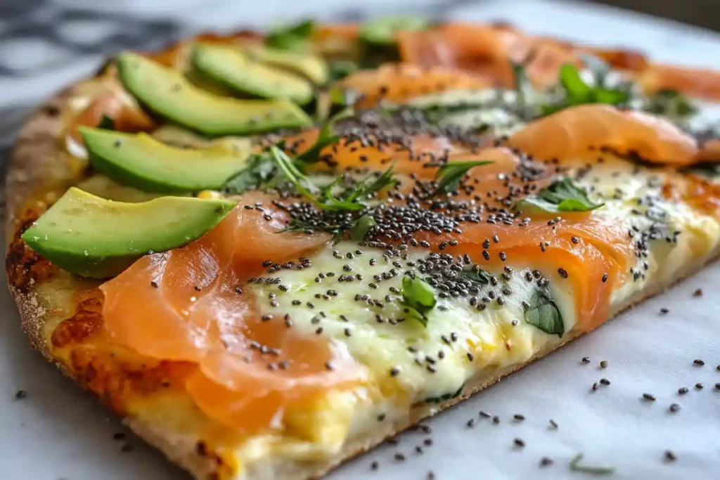 A close-up of a gourmet pizza topped with smoked salmon, avocado slices, melted cheese, chia seeds, and fresh herbs on a thin, crispy crust.