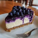 A close-up of a slice of fresh blueberry cheesecake with a golden graham cracker crust, creamy white cheesecake filling, and a glossy blueberry topping on a white plate with a fork beside it.