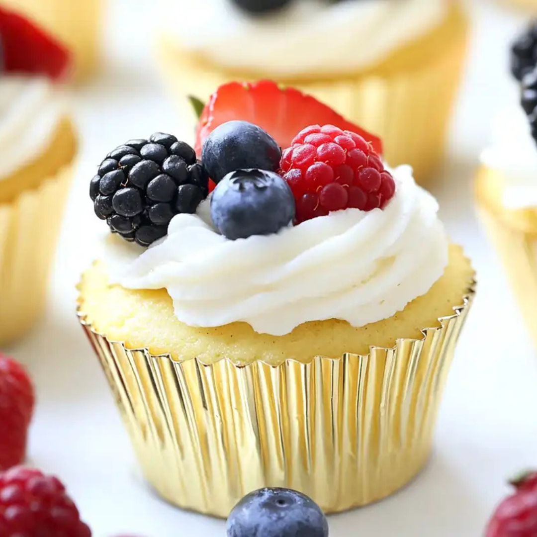 Mini cheesecake cupcakes in gold liners, topped with whipped cream and fresh blackberries, blueberries, raspberries, and strawberries.