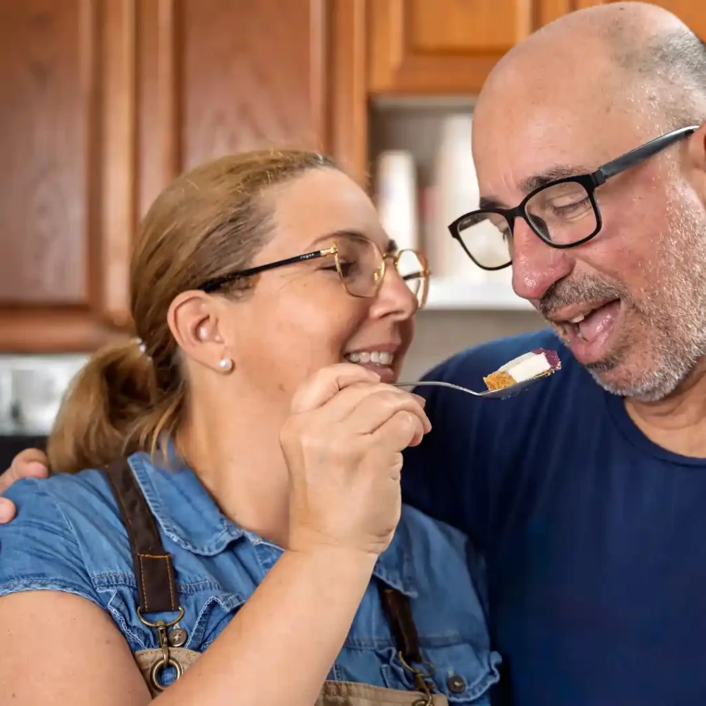 A smiling woman feeds a man a bite of dessert on a fork in a cozy kitchen.