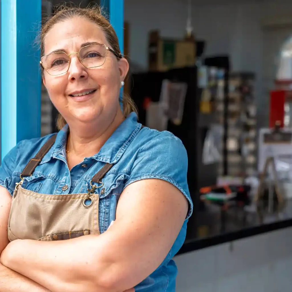 A confident woman wearing glasses and an apron stands with her arms crossed, smiling in front of a kitchen.