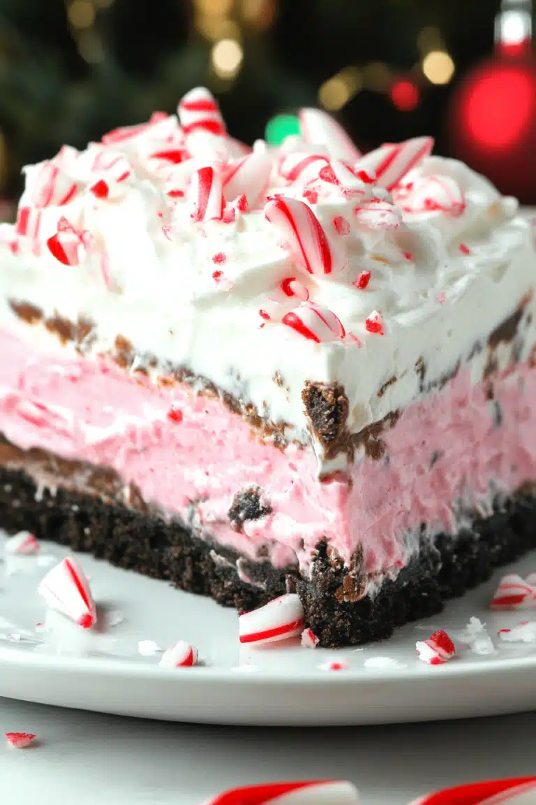 Close-up of a festive no-bake Christmas lasagna dessert slice with layers of chocolate crust, pink filling, and candy cane topping.